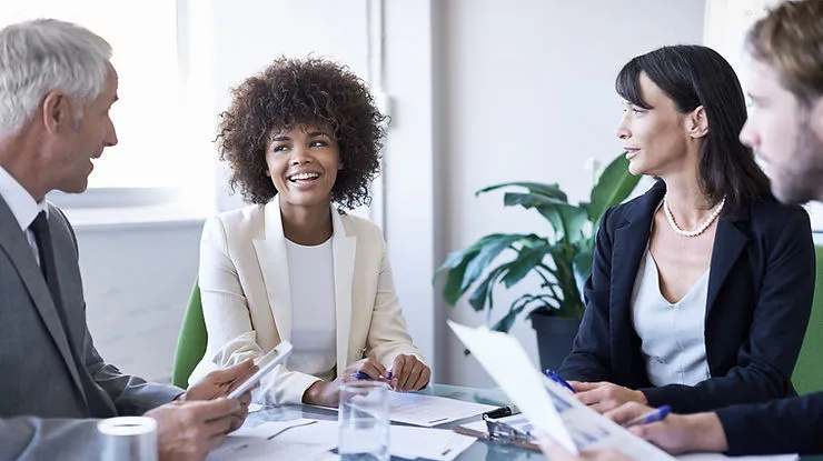 A group of diverse professionals sitting around a table in a bright office, engaged in a collaborative meeting.