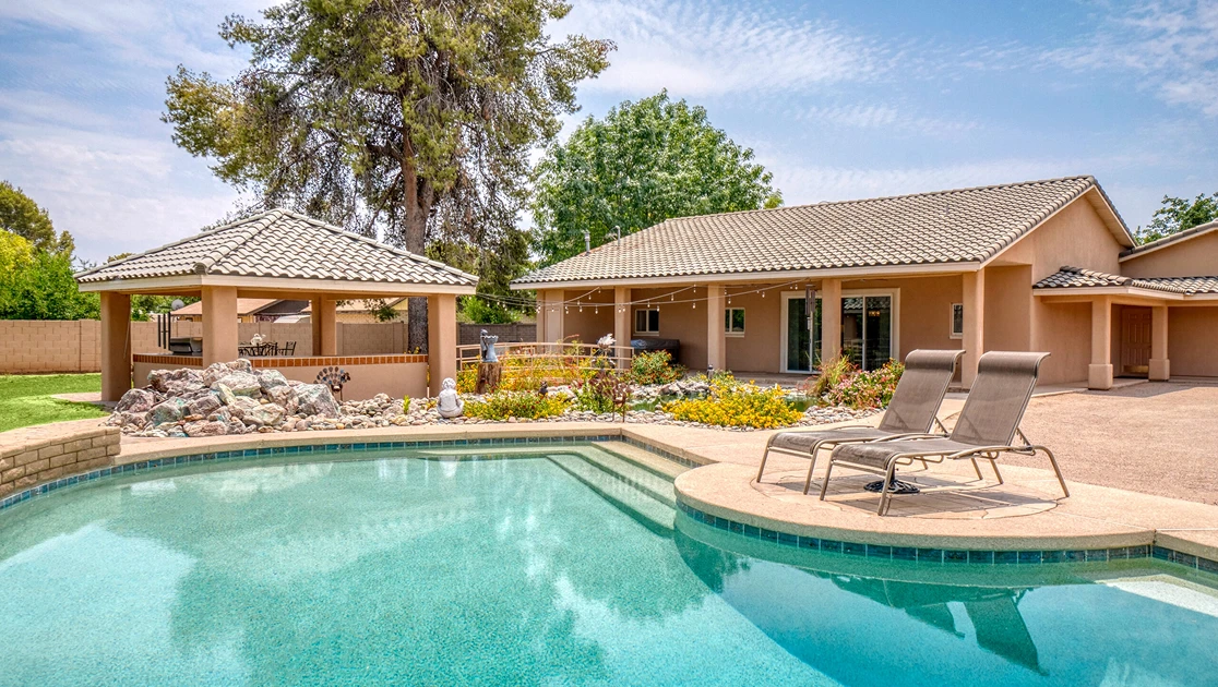 a backyard features a swimming pool with lounge chairs in the foreground, alongside a covered patio gazebo and a single-story house under a bright sky.