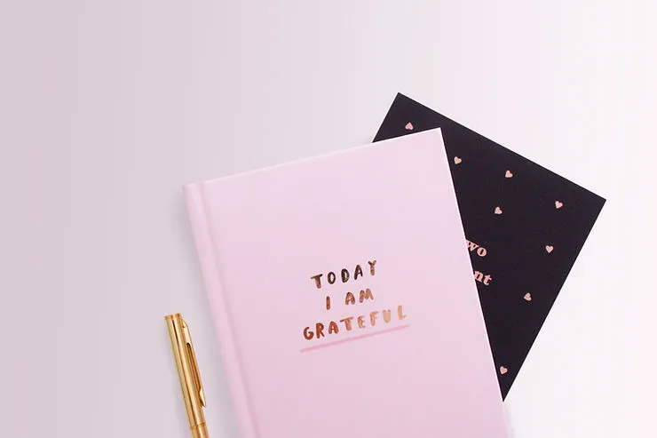 A light pink journal with the text "TODAY I AM GRATEFUL" in gold lettering sits on a desk next to a black card with a heart pattern and a gold pen in image